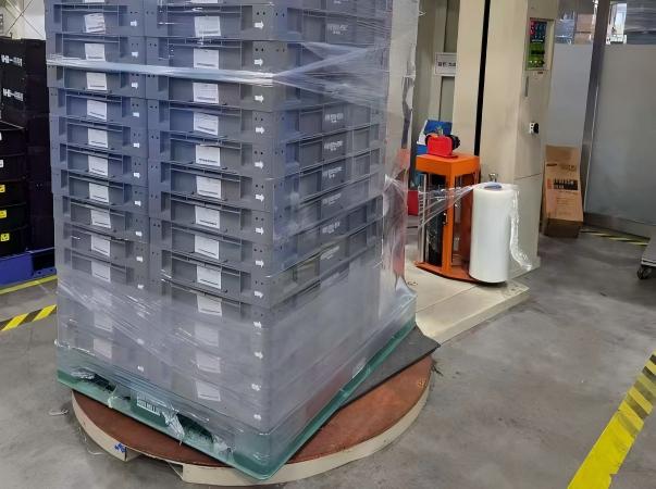 Uma fotografia industrial mostrando uma embaladora de paletes automatizada aplicando filme stretch de alto desempenho para m&aacute;quina em uma pilha alta de engradados de frutas. O processo de embalagem protege a carga agr&iacute;cola pesada para transporte, ao mesmo tempo que permite a circula&ccedil;&atilde;o de ar (ventila&ccedil;&atilde;o) necess&aacute;ria para manter o frescor do produto durante a log&iacute;stica e armazenamento da cadeia de frio.